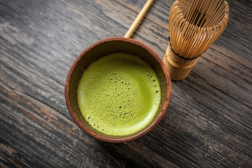 Braune Tasse Matcha-Tee auf holziger Oberfläche, mit einem Matcha-Besen.