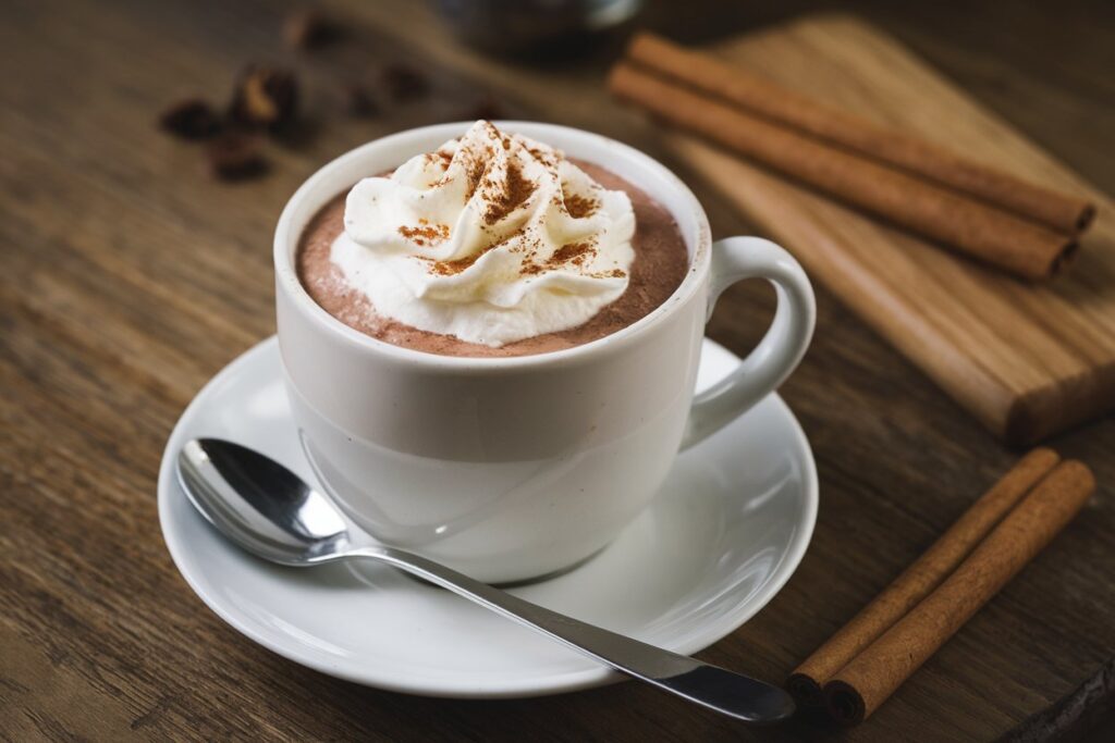 Eine Tasse Schokoladenmilch mit Zimt auf holziger Unterlage, Löffel und Zimtstangen im Hintergrund.